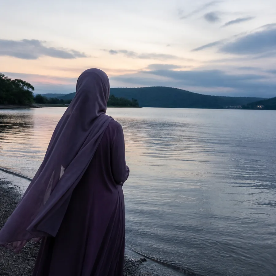A woman in a flowing dusty purple abaya and hijab standing at the edge of a calm lake at dusk, looking towards distant hills